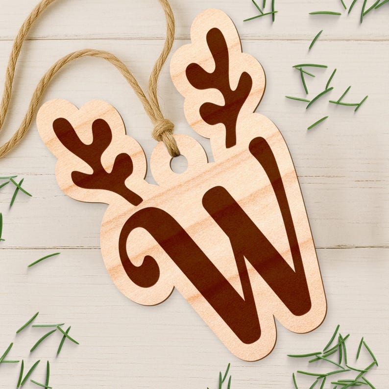 May include: Wooden Christmas ornament in the shape of a reindeer with the letter "W" in a dark brown color. The ornament is attached to a natural twine string. Green pine needles are scattered around the ornament on a white wooden surface.