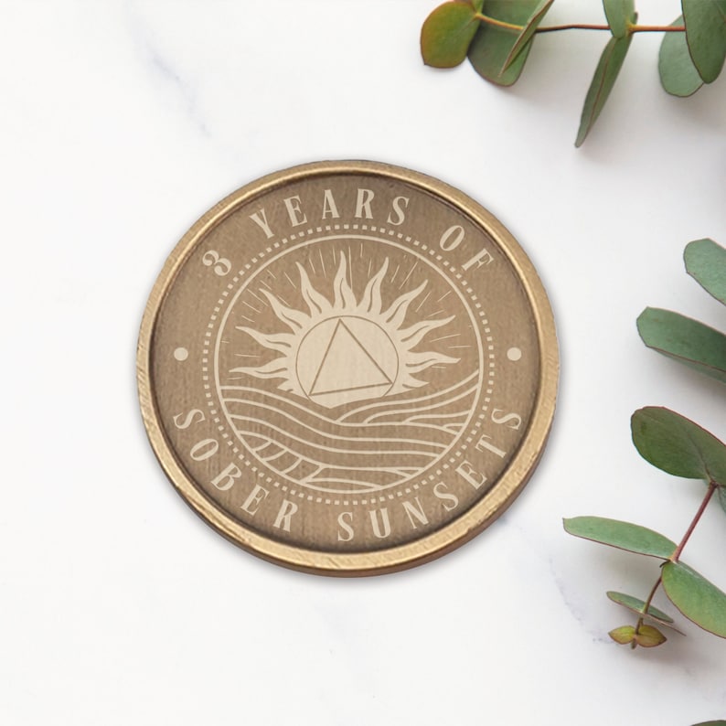 May include: A gold-toned round coin with the words "3 Years of Sober Sunsets" engraved around the edge. The center of the coin features a stylized sun with a triangle inside, and waves below.