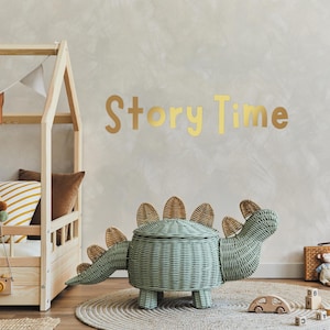 May include: A light green wicker dinosaur toy storage bin with a lid. The bin has brown wicker accents on the back and tail. The bin is sitting on a woven rug in front of a wooden bed with a gold "Story Time" sign on the wall.