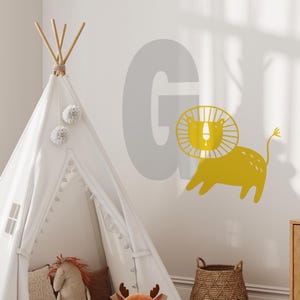 May include: A gray letter G with a yellow lion illustration. The lion has a mane and is standing on all fours. The image is on a white wall with a white teepee tent in the foreground.