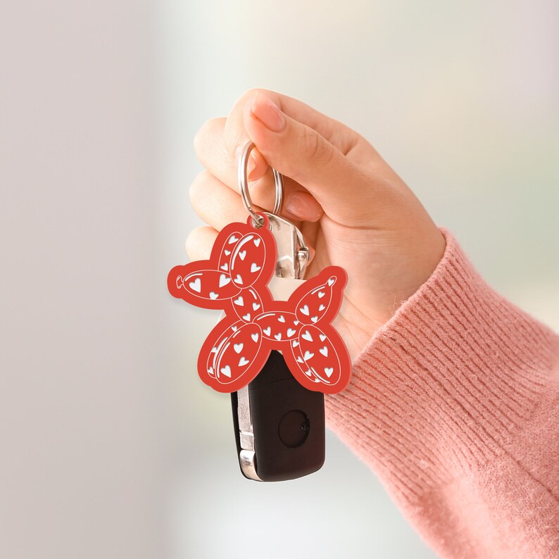 May include: A red keychain featuring a balloon dog design with white heart patterns. The keychain is attached to a silver key ring and a black car key. The background is blurred, with a person's hand holding the keychain.