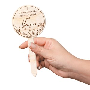 May include: A wooden sign with a round shape and a brown stick attached to the back. The sign has a white background and brown text that reads "If mum's were like flowers, I would pick You". The sign is decorated with a floral design.