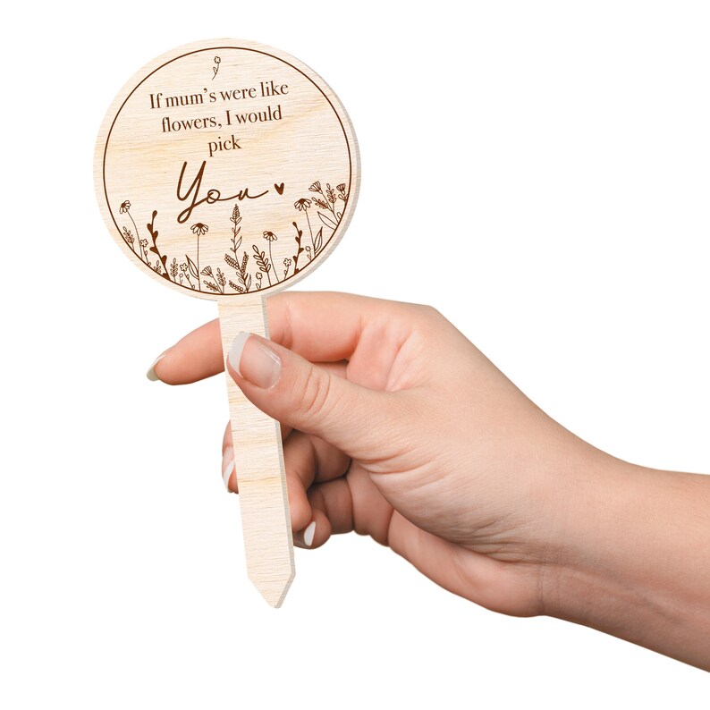 May include: A wooden sign with a round shape and a brown stick attached to the back. The sign has a white background and brown text that reads "If mum's were like flowers, I would pick You". The sign is decorated with a floral design.