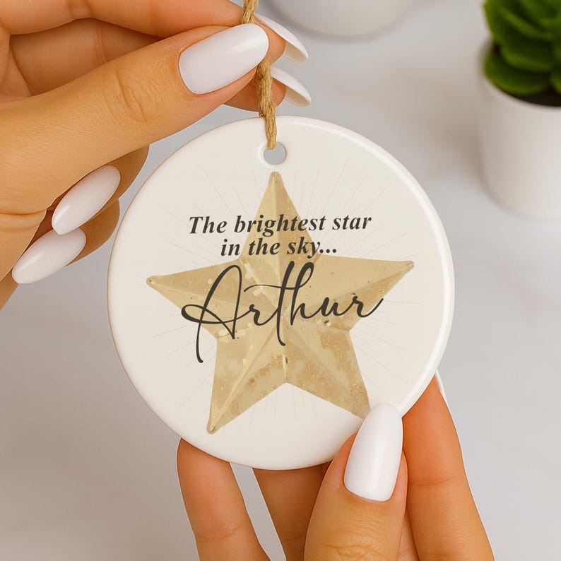 May include: A white ceramic ornament with a gold star design and the name "Arthur" in script. The text "The brightest star in the sky..." is above the name. A brown string is attached for hanging.