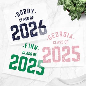May include: Three white squares with text for graduation announcements. The top square reads "-BOBBY- CLASS OF 2026" in navy blue glitter. The middle square reads "-FINN- CLASS OF 2025" in green. The bottom square reads "-GEORGIA- CLASS OF 2025" in pink.