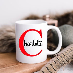 K&ouml;nnte beinhalten: Eine wei&szlig;e Keramiktasse mit einem roten Buchstaben "C" und dem Namen "Charlotte" in schwarzer Kursivschrift. Die Tasse steht auf einer Holzoberfl&auml;che.