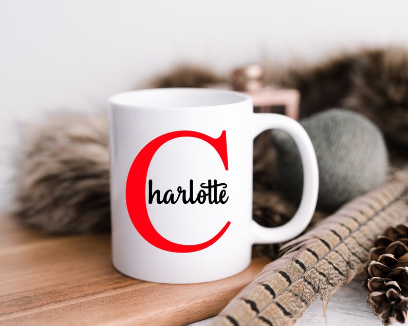 K&ouml;nnte beinhalten: Eine wei&szlig;e Keramiktasse mit einem roten Buchstaben "C" und dem Namen "Charlotte" in schwarzer Kursivschrift. Die Tasse steht auf einer Holzoberfl&auml;che.