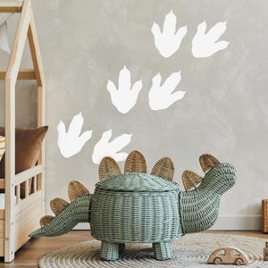 May include: A light green wicker dinosaur toy storage bin with a lid. The bin has a brown wicker tail and spines. The bin is sitting on a brown rug in front of a wall with white dinosaur footprints.