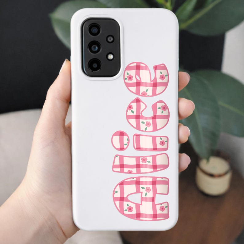 Puede incluir: Funda de tel&eacute;fono blanca con un m&oacute;dulo de c&aacute;mara y la palabra "NICE" en letras de cuadros rosas con peque&ntilde;os detalles florales. La funda se sostiene en una mano, con un fondo borroso de vegetaci&oacute;n y una peque&ntilde;a mesa de madera.