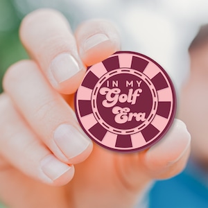 May include: A pink and maroon golf ball marker with the text "In My Golf Era" in a retro font.
