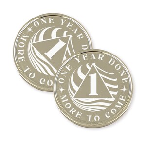 Op de afbeelding: Twee zilverkleurige metalen tokens met een wit ontwerp. De tokens hebben een driehoek met het nummer "1" erin. De tekst rond de driehoek luidt "One Year Done" en "One Year To Come".