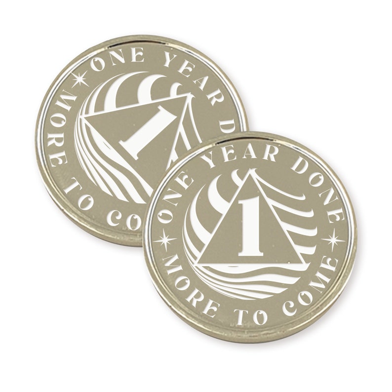 Op de afbeelding: Twee zilverkleurige metalen tokens met een wit ontwerp. De tokens hebben een driehoek met het nummer "1" erin. De tekst rond de driehoek luidt "One Year Done" en "One Year To Come".