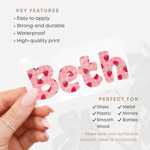 May include: A white sticker with the name "Beth" in pink letters, each decorated with red strawberries. The sticker is held by a hand with a gold ring. The sticker is perfect for glass, metal, plastic, mirrors, smooth surfaces, and bottles.