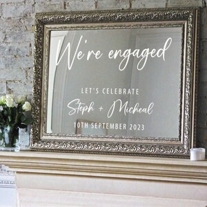 May include: A silver framed mirror with a white inscription that reads "We're engaged" with the text "Let's celebrate Steph + Micheal 10th September 2023" below.