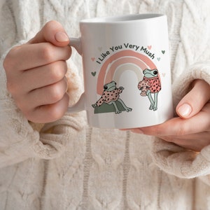 Puede incluir: Taza de cer&aacute;mica blanca con un arco&iacute;ris y dos ranas con su&eacute;teres. El texto en la taza dice "I Like You Very Mush".
