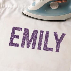 May include: A white shirt with the name "EMILY" in large, glittery purple letters. A blue and white iron is positioned above the letters, suggesting the application of the design to the fabric.