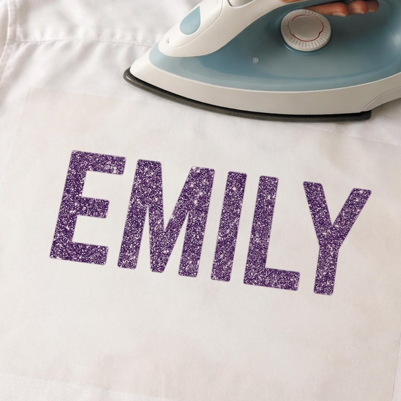 May include: A white shirt with the name "EMILY" in large, glittery purple letters. A blue and white iron is positioned above the letters, suggesting the application of the design to the fabric.
