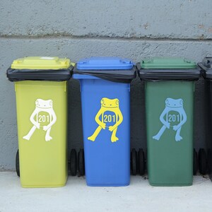 May include: A row of colorful recycling bins. The bins are yellow, blue, and green, each with a frog graphic holding a sign with the number 201. The bins have black lids and are lined up against a gray wall.