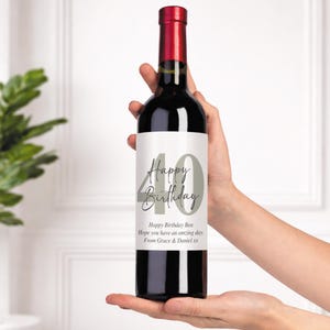 May include: A bottle of red wine with a label that reads "Happy Birthday" and the number "40" in gray. The label also includes a personalized message. The bottle has a red top and is held by a person.