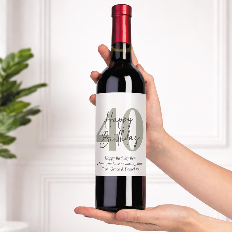 May include: A bottle of red wine with a label that reads "Happy Birthday" and the number "40" in gray. The label also includes a personalized message. The bottle has a red top and is held by a person.