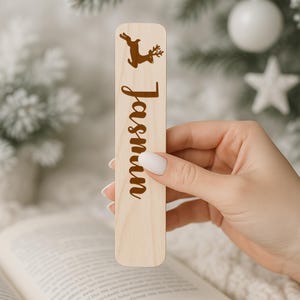 May include: A wooden bookmark with the name "Jasmin" in a brown script font. A brown reindeer silhouette is at the top. The bookmark is held over an open book, with a blurred background of Christmas decorations.