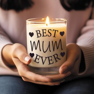 Op de afbeelding: Een transparante glazen kaars met een witte wasbasis en een zwarte inscriptie die "BEST MUM EVER" luidt met kleine hartjes aan weerszijden van de tekst. De kaars is aangestoken en de vlam is zichtbaar.