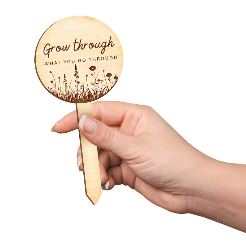 K&ouml;nnte beinhalten: Ein h&ouml;lzerner Gartenpfahl mit einem runden Schild, auf dem "Grow through what you go through" steht. Das Schild hat eine Silhouette von Blumen und Gras.