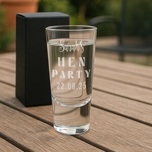 Op de afbeelding: Een helder shotglaasje met de witte tekst "Sarah's HEN PARTY 22.08.25", gevuld met vloeistof. Achter het glas staat een zwarte geschenkdoos, op een houten oppervlak.