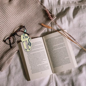 May include: A close-up of an open book with a cat-shaped bookmark. The bookmark is yellow with black markings and is placed between the pages of the book. The book is on a white, textured surface.
