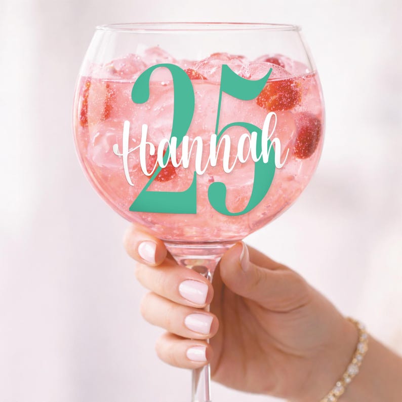 May include: A large, clear glass of pink cocktail with ice and strawberries. The glass has the number "25" in teal and the name "Hannah" in white script. The glass is held by a person.