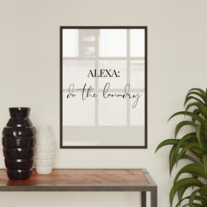 May include: A black framed print with the text "ALEXA: do the laundry" in white lettering on a white background.