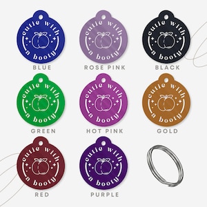 May include: A selection of pet tags in various colours, each featuring the text "cutie with a booty" and a peach illustration. The tags are in blue, rose pink, black, green, hot pink, gold, red, and purple. A silver split ring is also included.