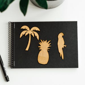 May include: Three wooden cutouts on a black background. The cutouts are a palm tree, a pineapple, and a parrot. The cutouts are a light brown color.