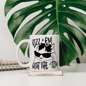 May include: White ceramic mug with a black and white illustration of a cat wearing sunglasses. The text "Rizz 'em with the 'tism" is printed on the mug.