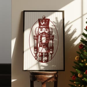 May include: A framed print featuring a detailed illustration of a traditional nutcracker soldier in burgundy against a cream background with vertical stripes. The nutcracker is inside an oval frame. The print is displayed on a wooden table.
