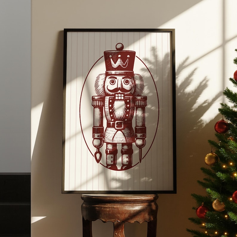 May include: A framed print featuring a detailed illustration of a traditional nutcracker soldier in burgundy against a cream background with vertical stripes. The nutcracker is inside an oval frame. The print is displayed on a wooden table.