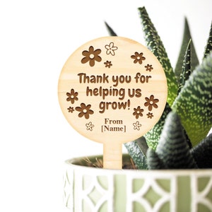 May include: Wooden plant stake with a round, brown, engraved sign that says "Thank you for helping us grow! From [Name]" with a flower design around the text.