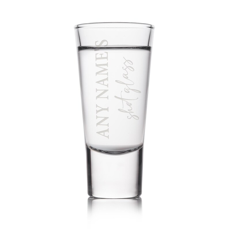 May include: A clear shot glass with the text "ANY NAME'S shot glass" etched on the front. The glass is filled with a clear liquid. The shot glass has a thick base and a straight, cylindrical body.