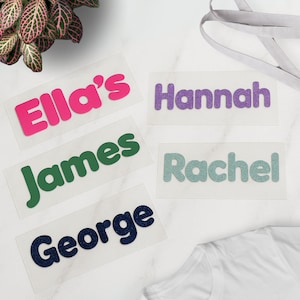 May include: Five iron-on name labels with different names. The names are Ella's, Hannah, James, Rachel, and George. The labels are on a white surface.