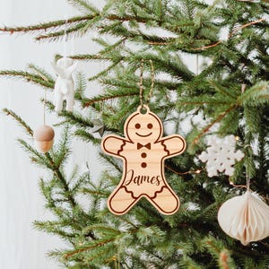 Puede incluir: Un adorno navide&ntilde;o de madera en forma de hombre de jengibre con el nombre "James" grabado en &eacute;l. El adorno est&aacute; colgado en un &aacute;rbol de Navidad.