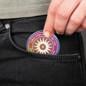Op de afbeelding: Een ronde, veelkleurige soberheidsmunt met de tekst "sober trips around the sun" en een zonnesterontwerp met het nummer "6" in het midden. De munt wordt in een zwarte jeanszak gehouden.