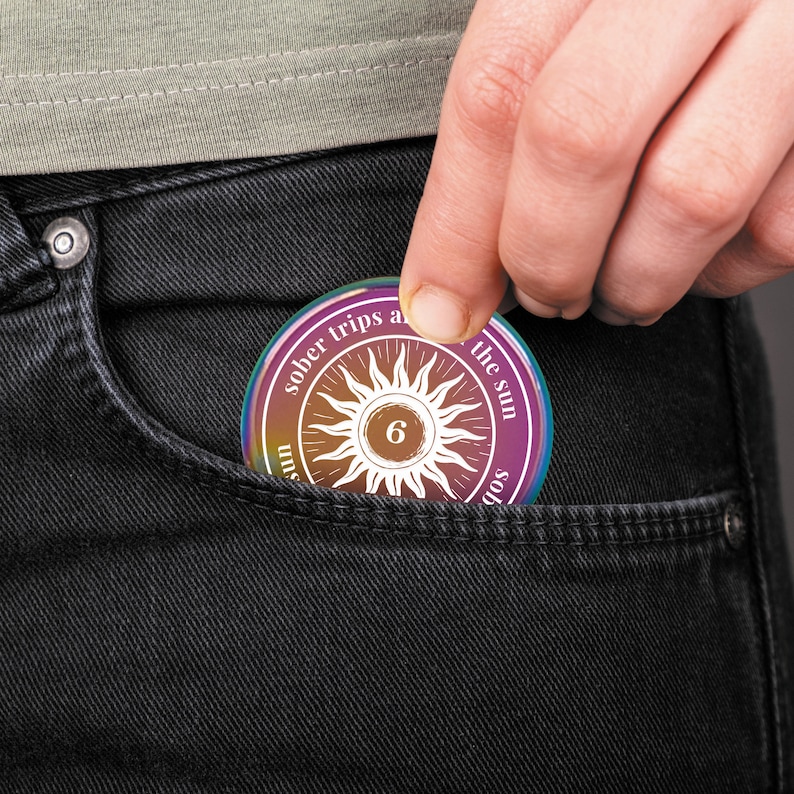 Op de afbeelding: Een ronde, veelkleurige soberheidsmunt met de tekst "sober trips around the sun" en een zonnesterontwerp met het nummer "6" in het midden. De munt wordt in een zwarte jeanszak gehouden.