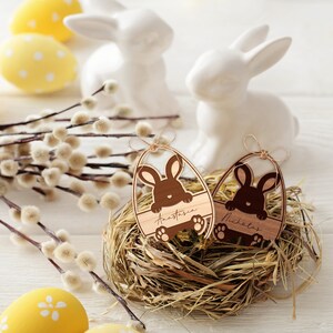 May include: Two wooden Easter egg ornaments with bunny cutouts and personalized names "Anastasia" and "Nicholas". The ornaments are nestled in a straw nest.