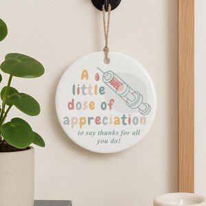 May include: A round, white ceramic hanging decoration with the text "A little dose of appreciation" and a syringe illustration. The text is in pastel colors, and the syringe is red and gray. A jute string is attached for hanging.