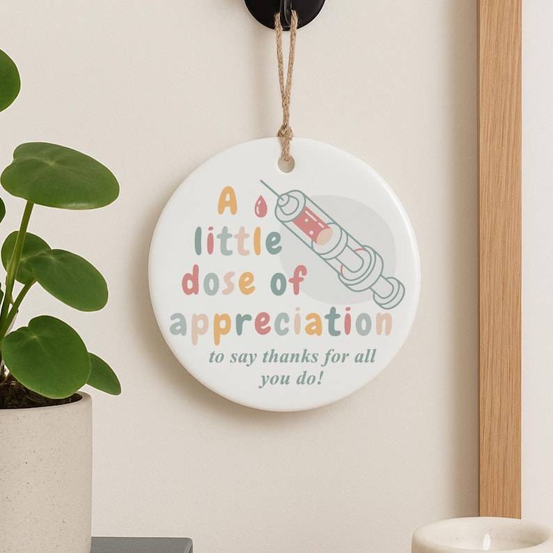 May include: A round, white ceramic hanging decoration with the text "A little dose of appreciation" and a syringe illustration. The text is in pastel colors, and the syringe is red and gray. A jute string is attached for hanging.