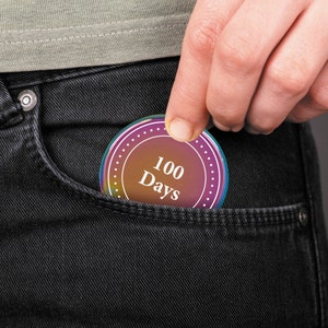 May include: A round, iridescent, holographic sticker with a white border and white text that reads "100 Days" is placed in a dark blue jean pocket.