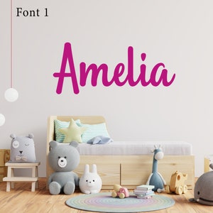 Op de afbeelding: Een houten bed met een grijze teddybeer en ander speelgoed, met de naam "Amelia" in roze cursief op een witte muur.