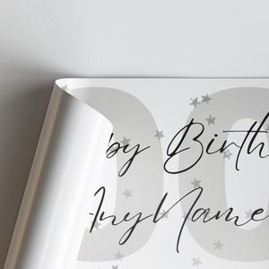 May include: A partially visible, rolled-up poster with a gray background and large gray numbers. The poster features the text "by Birth" and "Any Name" in elegant black script, along with scattered star shapes.