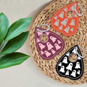 May include: Three different colored keychains shaped like a ghost with a hole in the middle. The keychains are on a woven straw mat. The ghosts are red with white ghosts, purple with pink ghosts, and black with white ghosts.