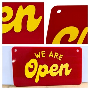 May include: A rectangular red sign with the words "WE ARE Open" in yellow, stylized lettering. The sign has rounded corners and two holes for hanging. The background is white.